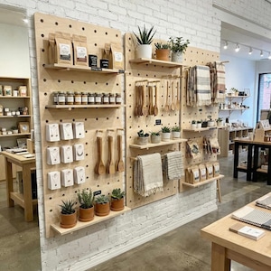 以下が含まれることがあります： さまざまな商品が展示された木製のペグボードの小売ディスプレイ。パッケージ商品、ジュエリー、木製食器、鉢植え、テキスタイルなど。白いレンガの壁とコンクリートの床に設置されています。