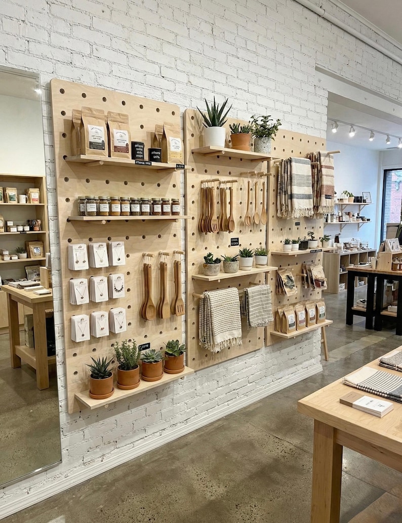 以下が含まれることがあります： さまざまな商品が展示された木製のペグボードの小売ディスプレイ。パッケージ商品、ジュエリー、木製食器、鉢植え、テキスタイルなど。白いレンガの壁とコンクリートの床に設置されています。