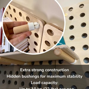 Op de afbeelding: Een lichtgekleurd houten pegboard-wandsysteem met cilindrische houten pinnen en witte bussen. De tekst luidt: Extra strong construction, Hidden bushings for maximum stability, Load capacity: Up to 10 kg (22 lbs) per peg, Up to 20 kg (44 lbs) per shelf.