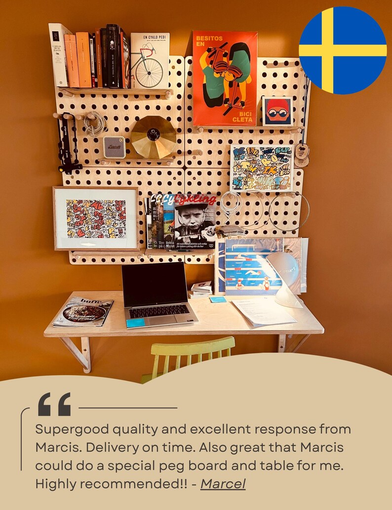 May include: A light-colored wooden pegboard wall organizer system mounted above a matching wooden desk. The pegboard features various shelves holding books, a gold disc, a small speaker, framed art, and magazines. A laptop sits on the desk.