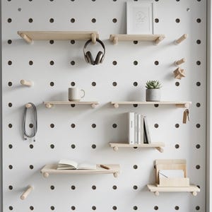 May include: A white pegboard with wooden shelves and pegs, displaying various items. These include a mug, headphones, books, a framed picture, a succulent, and a notebook. The pegboard is mounted on a white wall. The shelves are made of light-colored wood.