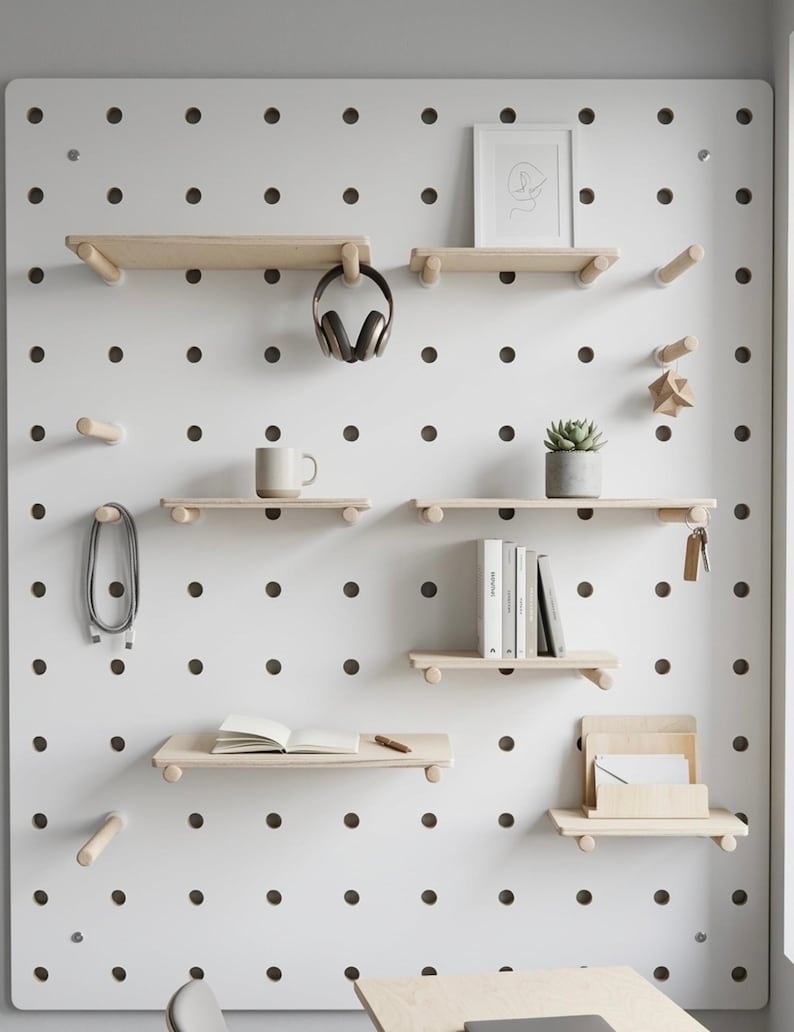 May include: A white pegboard with wooden shelves and pegs, displaying various items. These include a mug, headphones, books, a framed picture, a succulent, and a notebook. The pegboard is mounted on a white wall. The shelves are made of light-colored wood.