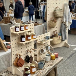 以下が含まれることがあります： 屋外市場の手作り品の展示。木製の棚には、ジャムの瓶、革のポーチ、マグカップ、キャンドル、籐のバスケットが並んでいます。素朴な木製のテーブルとペグボードのスタンドに商品が配置されています。