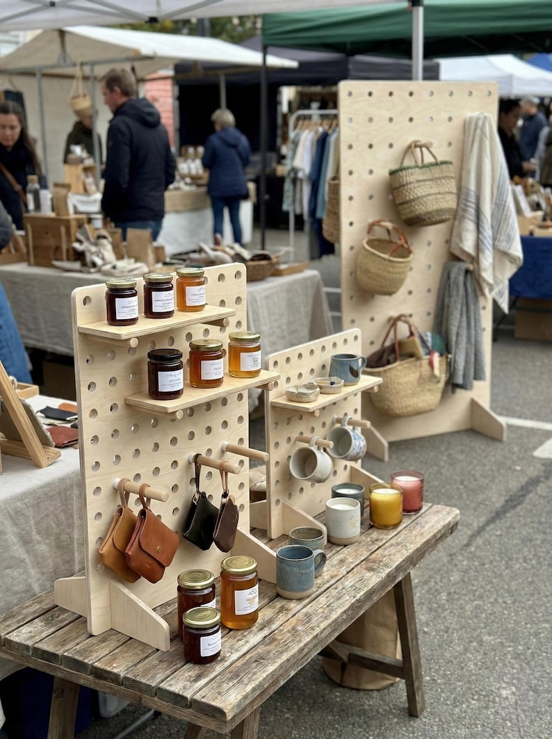 以下が含まれることがあります： 屋外市場の手作り品の展示。木製の棚には、ジャムの瓶、革のポーチ、マグカップ、キャンドル、籐のバスケットが並んでいます。素朴な木製のテーブルとペグボードのスタンドに商品が配置されています。