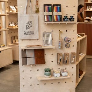 May include: A wooden display stand showcasing stationery products. The display includes colored pencils, ink bottles, notebooks, pens, washi tape, and a tote bag with a leaf logo. The stand is on wheels and has a sign that reads "Rado&scaron;ais Stūritis".