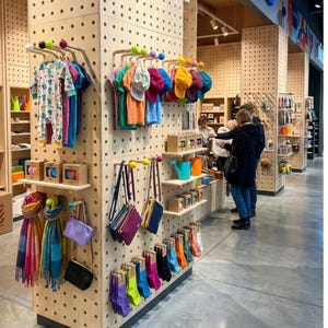 Op de afbeelding: Een winkeldisplay van lichtgekleurde geperforeerde houten panelen met diverse artikelen zoals kleurrijke kleding, hoeden, sjaals, kleine handtassen en sokken die aan houten haringen met felgekleurde bolvormige uiteinden hangen.