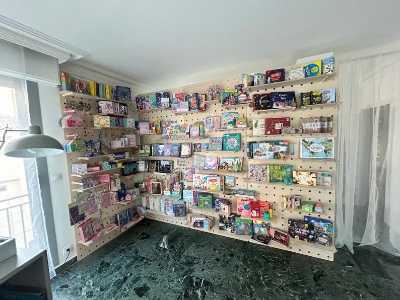 May include: A display of various books and small items arranged on wooden shelves attached to a pegboard wall. The items include books of different sizes, colors, and designs, along with other decorative items. The wall is light-colored, and the shelves are evenly spaced.
