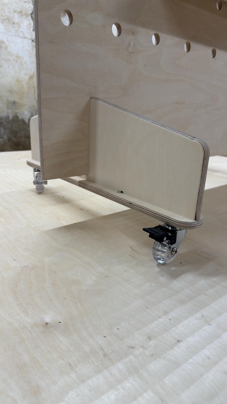 May include: A light-coloured wooden storage unit with a series of evenly spaced, semi-circular cutouts along the top edge. The unit has a side panel with a smaller, angled section and is mounted on clear caster wheels.