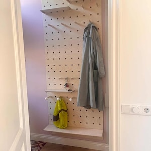 May include: A wooden pegboard with shelves and pegs, designed for entryway organization. A gray coat and a yellow bag hang from the board. The pegboard is mounted on a light pink wall.