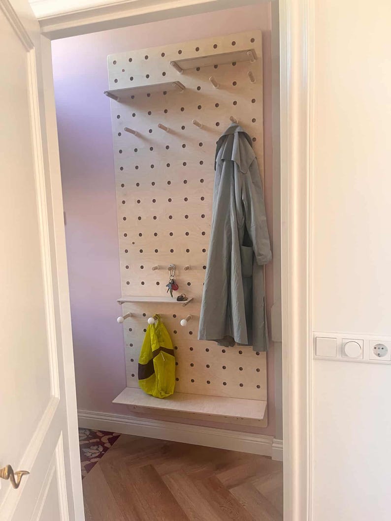 May include: A wooden pegboard with shelves and pegs, designed for entryway organization. A gray coat and a yellow bag hang from the board. The pegboard is mounted on a light pink wall.