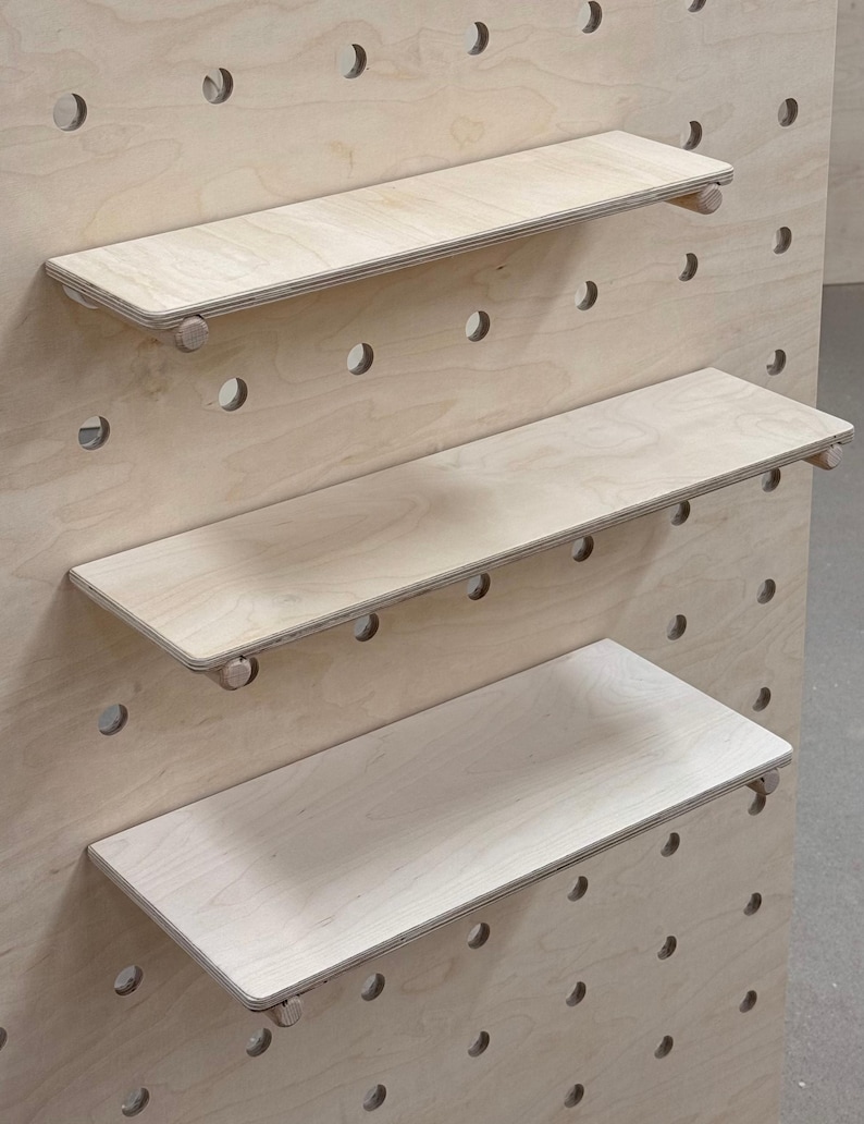 May include: A light-colored wooden pegboard with three shelves. The shelves are rectangular and supported by wooden dowels. The pegboard has evenly spaced holes for shelf placement. The wood has a natural grain.