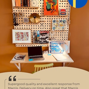 May include: A light-colored wooden pegboard wall organizer system mounted above a matching wooden desk. The pegboard features various shelves holding books, a gold disc, a small speaker, framed art, and magazines. A laptop sits on the desk.