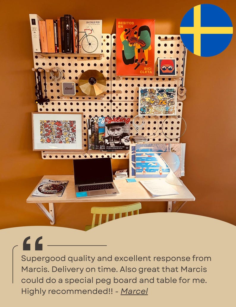 May include: A light-colored wooden pegboard wall organizer system mounted above a matching wooden desk. The pegboard features various shelves holding books, a gold disc, a small speaker, framed art, and magazines. A laptop sits on the desk.