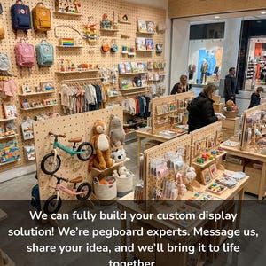 Op de afbeelding: Een winkelinterieur met lichtgekleurde houten pegboard-wanden en vrijstaande displays. De schappen bevatten diverse kinderartikelen, waaronder rugzakken, knuffels, loopfietsen, boeken en kleding aan kledinghangers.