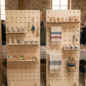 May include: Two wooden display stands with shelves and pegboard holes showcase various handcrafted items. The stands hold ceramic mugs, bowls, wooden toys, and woven textiles. The items are arranged on shelves and hung from pegs.