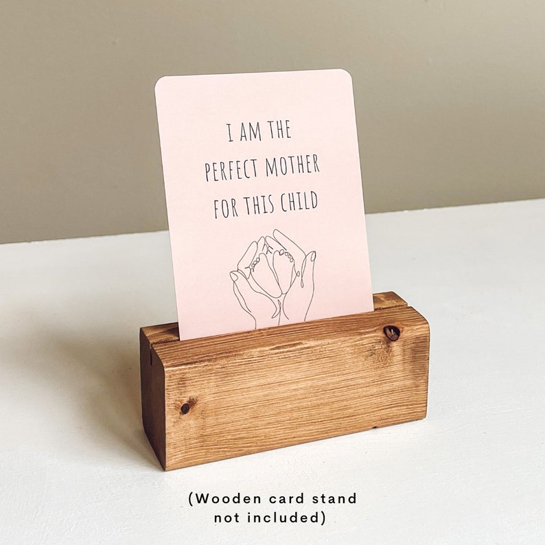 K&ouml;nnte beinhalten: Rosa Karte mit dem Text "Ich bin die perfekte Mutter f&uuml;r dieses Kind" und einer Strichzeichnung von H&auml;nden, die die F&uuml;&szlig;e eines Babys halten. Die Karte wird auf einem Holzst&auml;nder pr&auml;sentiert.