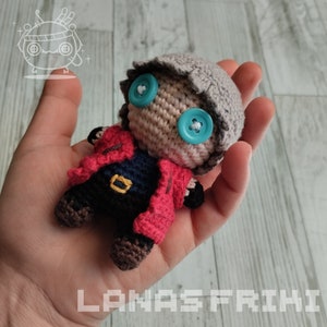 Puede incluir: Muñeca de ganchillo hecha a mano con cabeza gris, ojos de botón azules y chaqueta roja. La muñeca tiene un cuerpo negro con hebilla dorada y botas marrones. La muñeca se sostiene en una mano.