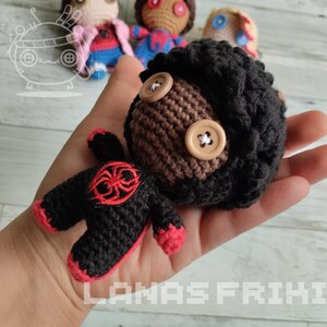 May include: A handmade crochet doll with a black body, red accents, and a brown head with black hair. The doll has button eyes and is held in a hand. Other dolls are visible in the background.