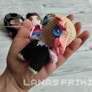 May include: A collection of handmade crochet dolls, each with unique features. One doll is held in a hand, showcasing its beige head with blue button eyes and pink accents. Other dolls are visible in the background. The dolls have black and white clothing.