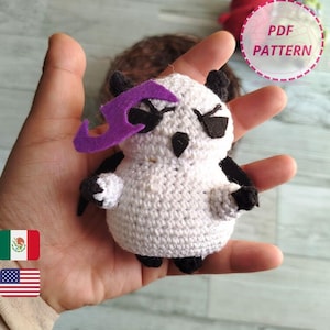 Op de afbeelding: Een handgemaakte witte gehaakte uil met zwarte accenten, een paars vilten accessoire en zwarte ogen. De uil houdt een klein zwart voorwerp vast. De afbeelding bevat de tekst "PDF PATTERN".