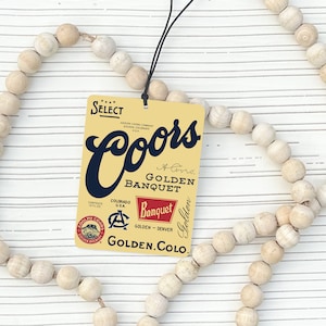 May include: A rectangular tag featuring the Coors Golden Banquet logo in navy blue and red on a yellow background, suspended by a black string. The tag is set against a backdrop of a beaded garland.