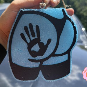 May include: A light blue, textured, butt-shaped hanging ornament featuring a black graphic of a handprint centered on the glute area. The design includes black outlines and black accents at the bottom, with a small metal loop at the top for hanging.
