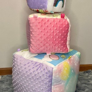 Peut inclure: Une pile de trois jouets pour bébés carrés, doux et colorés. Chaque jouet a un motif et une texture différents. Le jouet du haut a un côté blanc avec un motif arc-en-ciel, le jouet du milieu a un côté rose avec une surface texturée et le jouet du bas a un côté violet avec une surface texturée.