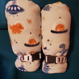 May include: A pair of white fleece arm warmers with a space-themed print. The warmers have a black and white adjustable strap with a buckle.