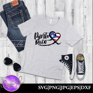 May include: A gray t-shirt with the words "Puerto Rico" in black lettering and a heart with the Puerto Rican flag inside. The shirt is on a wooden surface with a pair of jeans, sunglasses, and black high-top sneakers.