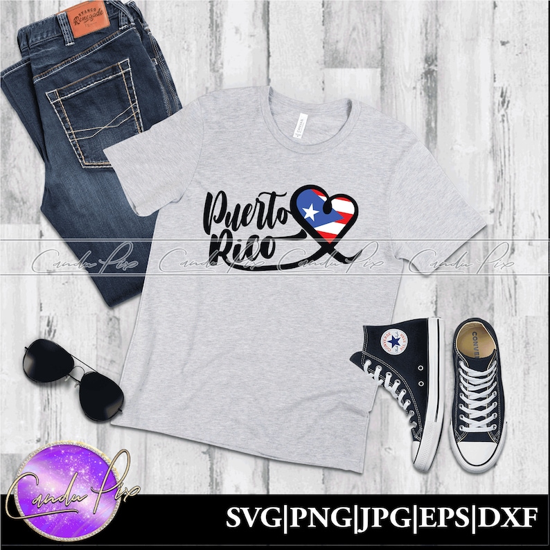 May include: A gray t-shirt with the words "Puerto Rico" in black lettering and a heart with the Puerto Rican flag inside. The shirt is on a wooden surface with a pair of jeans, sunglasses, and black high-top sneakers.
