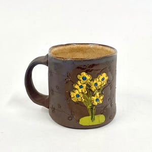 May include: A brown ceramic mug with a handle, featuring a hand-painted design of yellow flowers with blue centers on a brown background.