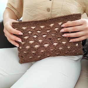 May include: A brown crocheted clutch bag with a geometric pattern. The bag has a woven texture and is held by a person's hands.