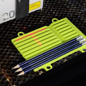 Könnte beinhalten: Ein grüner Plastik-Bleistifthalter mit einem Laser-Schnittmuster und drei blauen Bleistiften. Der Halter hat eine Schlaufe oben und steht auf einer schwarzen Wabenoberfläche.