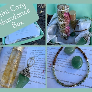 May include: A Mini Cozy Abundance Box, featuring a sage bundle, essential oil, a necklace with green beads, and a green crystal pendant. The box also includes a journal and a clear bottle with yellow liquid.