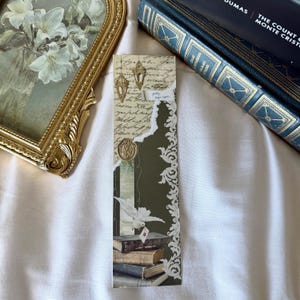 May include: A rectangular paper bookmark featuring a collage design with a beige background of cursive script, gold lantern illustrations, a white dove carrying a letter, and a stack of books. The bookmark includes a dark green section with white filigree.