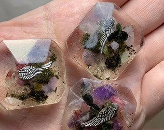 May include: Three clear resin pendants with a silver wing charm, filled with colorful stones and sand. The pendants are in a geometric shape, showcasing a unique, handcrafted design.
