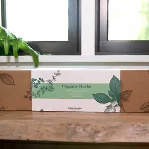 May include: A rectangular cardboard box labeled "Organic Herbs" with a green and white label. The box features botanical illustrations of herbs and the text "Home garden seed kit". The box is on a wooden surface.