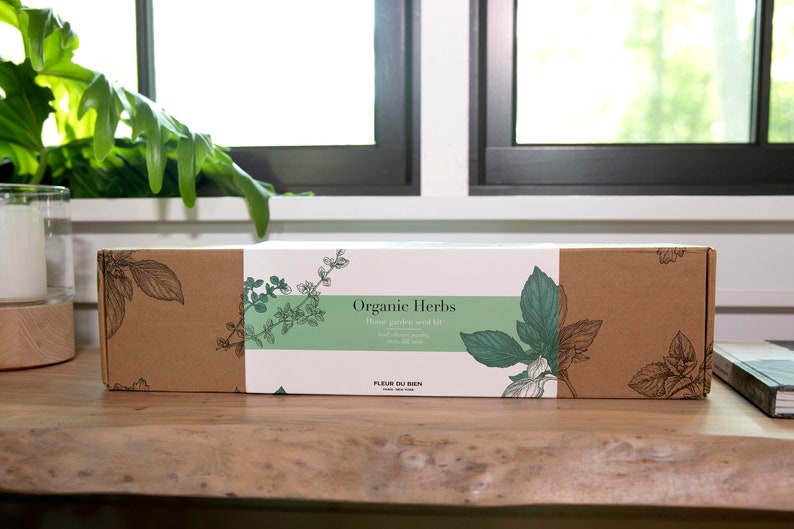 May include: A rectangular cardboard box labeled "Organic Herbs" with a green and white label. The box features botanical illustrations of herbs and the text "Home garden seed kit". The box is on a wooden surface.