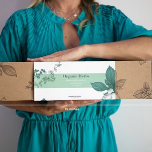 May include: A rectangular brown box, 19 inches long and 4.5 inches high, with a white label that reads "Organic Herbs". The label has botanical illustrations and the text "Home garden seed kit". The box is held by a person wearing a teal dress.