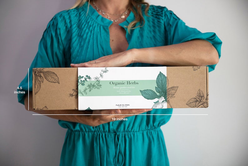 May include: A rectangular brown box, 19 inches long and 4.5 inches high, with a white label that reads "Organic Herbs". The label has botanical illustrations and the text "Home garden seed kit". The box is held by a person wearing a teal dress.