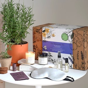 Op de afbeelding: Een witte tafel toont een zelfzorgset met een doos met het label "The Harmony Set". De set bevat een kaars, slaapmasker, essentiële oliën en een lavendelplant in een oranje pot. De doos heeft bloemenillustraties.