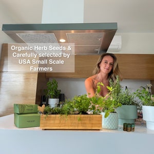 May include: A kitchen scene featuring a woman surrounded by potted herbs and a wooden planter box filled with various green herbs. The text "Organic Herb Seeds & Carefully selected by USA Small Scale Farmers" is visible.