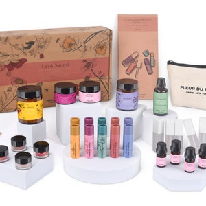 Könnte beinhalten: Eine Sammlung von Lippenpflegeprodukten, darunter Tiegel, Lipgloss und ätherische Öle, die auf weißen Podesten angeordnet sind. Die Produkte sind in verschiedenen Farben verpackt und mit der Marke "Fleur du Bien" versehen. Eine Canvas-Tasche mit dem Markennamen ist ebenfalls enthalten.