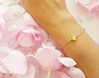 May include: A delicate gold bracelet with a small hexagonal charm featuring the letter "H". The bracelet is shown on a wrist against a background of pink fabric flowers.