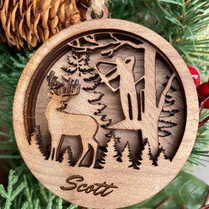 May include: A wooden Christmas ornament with a deer and a hunter in a forest scene. The ornament is personalized with the name "Scott".