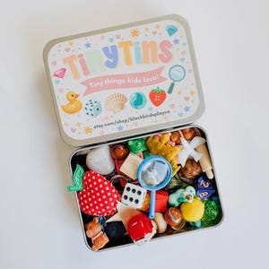 May include: An open metal tin labeled "Tiny Tins" filled with small toys and trinkets. The lid features a colorful design with a duck, dice, seashell, and strawberry. Inside are a magnifying glass, dice, and a red strawberry.
