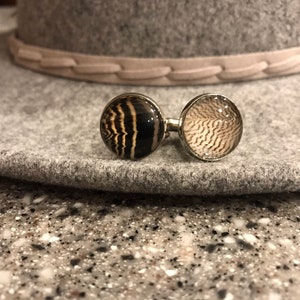 May include: A pair of cufflinks with a silver metal base and round glass tops. One cufflink features a black and white striped pattern, while the other has a brown and white feathered pattern.
