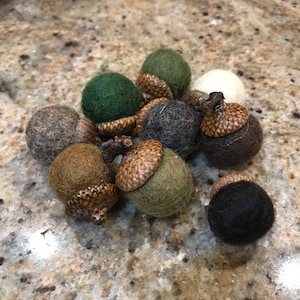May include: A collection of felt acorns in various shades of green, brown, and gray. The acorns have a realistic appearance with brown felt caps.