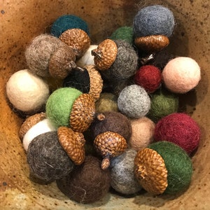 May include: A collection of handmade felt acorns in a variety of colors, including green, brown, gray, and burgundy. The acorns are nestled in a rustic, speckled ceramic bowl, creating a warm, autumnal aesthetic.