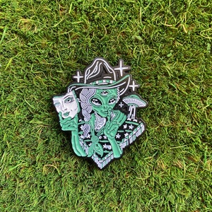Op de afbeelding: Groene emaille pin met een alien die een heksenhoed draagt en een wit masker vasthoudt. De alien heeft drie ogen en draagt een gepatroneerde sjaal. De pin heeft een zwarte omtrek en is geplaatst tegen een groene achtergrond.
